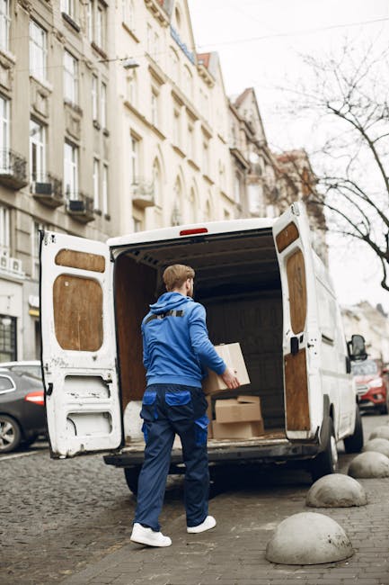 A man dressed in a blue jacket, dark trousers, and white shoes is standing on a cobblestone street near a white delivery van with its rear doors open. Inside the van, visible wooden paneling and some cardboard boxes indicate a home relocation process. The man is holding a cardboard box, preparing to load it onto the van. The surrounding environment includes historic multi-storey buildings with ornate facades, parked cars along the street, and a leafless tree, suggesting a cold season. Large concrete bollards are positioned in front of the van to prevent accidental vehicle overrun. The scene captures a furniture transport or packing process, with the van likely being used for moving services as provided by Man and a Van Bayswater, facilitated for house removals in the W2 area.