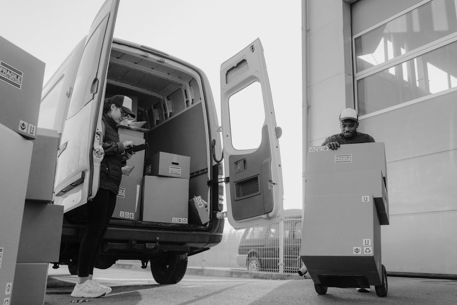In this black-and-white photograph, two individuals are engaged in a home relocation process involving furniture transport and packing materials outside a modern building. The woman on the left, wearing a cap, glasses, and a padded jacket, stands inside the open rear doors of a large delivery van, which is filled with cardboard boxes, some wrapped in plastic, and other packing supplies stacked on shelves. She appears to be preparing or organizing items for loading or unloading as part of a professional removals service. The man on the right, dressed in a cap and dark clothing, is operating a wheeled trolley with a sizable cardboard box on it, which is branded with the company's labels. He is positioned on the pavement just outside the building, next to the loading bay area with a large window and smooth exterior wall visible in the background. The scene captures the essential steps of a furniture transfer and loading process carried out by Man and a Van Bayswater, emphasizing efficient logistics in house removals.