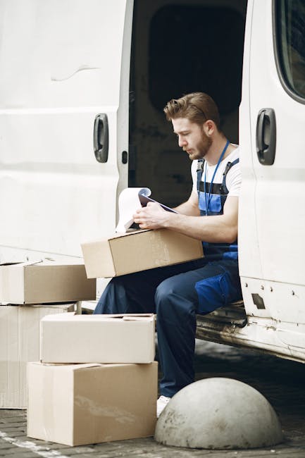 A man dressed in a blue jacket, dark trousers, and white shoes is standing on a cobblestone street near a white delivery van with its rear doors open. Inside the van, visible wooden paneling and some cardboard boxes indicate a home relocation process. The man is holding a cardboard box, preparing to load it onto the van. The surrounding environment includes historic multi-storey buildings with ornate facades, parked cars along the street, and a leafless tree, suggesting a cold season. Large concrete bollards are positioned in front of the van to prevent accidental vehicle overrun. The scene captures a furniture transport or packing process, with the van likely being used for moving services as provided by Man and a Van Bayswater, facilitated for house removals in the W2 area.