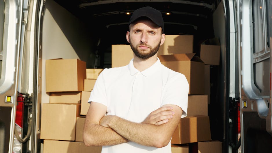Two individuals are engaged in a home relocation process, working inside the open rear of a large white van positioned on the pavement outside a residential property. The man, dressed in a white polo